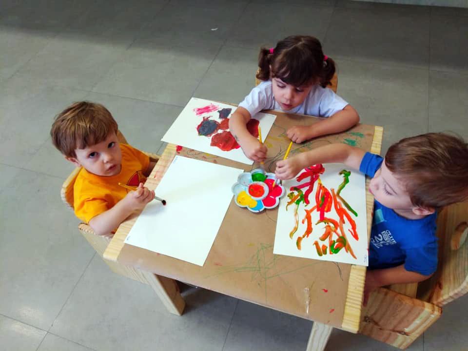 Três crianças pintando com tinta guache em uma mesinha, cada uma com sua folha e pincel