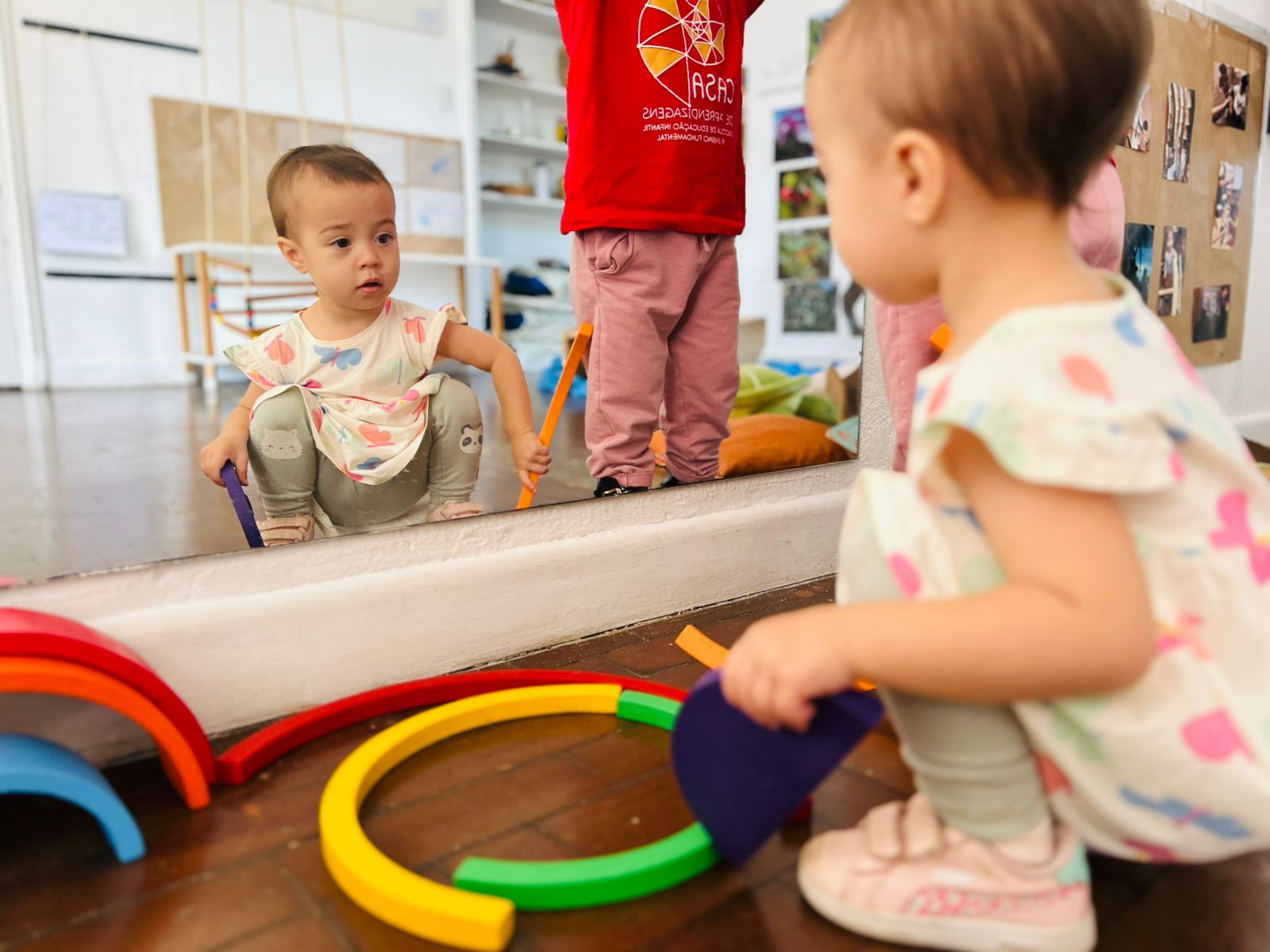 Bebê brincando com arco-íris de madeira em frente ao espelho, com educadora ao fundo